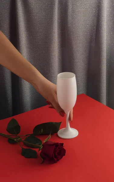 Kadın eli, şarap kadehi ve kırmızı gül çiçekli en az romantik kompozisyon. Parlak gri perde arkaplanı. Retro Sevgililer Günü partisi konsepti.