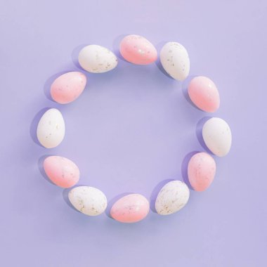 Mor arka planda pembe pastel renklerle boyanmış renkli yumurtalardan yapılmış yaratıcı Paskalya çelengi. Düz yatıyordu. Boşluğu kopyala.