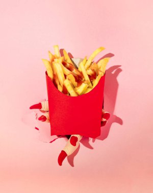 Creative layout made of hand with red nails holding french fries in red paper box against pastel pink background. Halloween surreal spooky idea.
