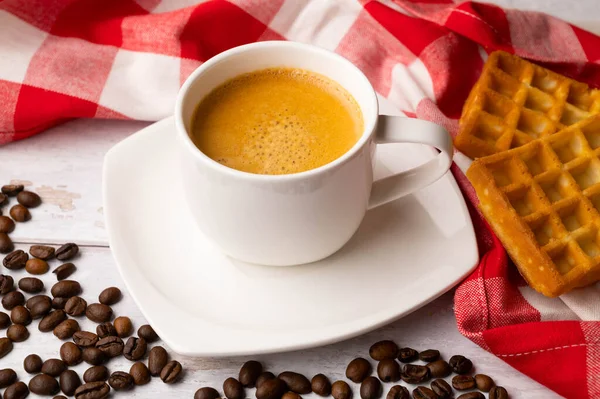 A cup of coffee with Belgian waffles and spilled coffee beans on a white wooden table. Around a red and white checkered kitchen towel.