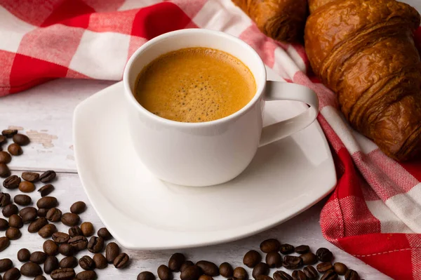 A cup of coffee with croissants and spilled coffee beans on a white wooden table. Around a red and white checkered kitchen towel.