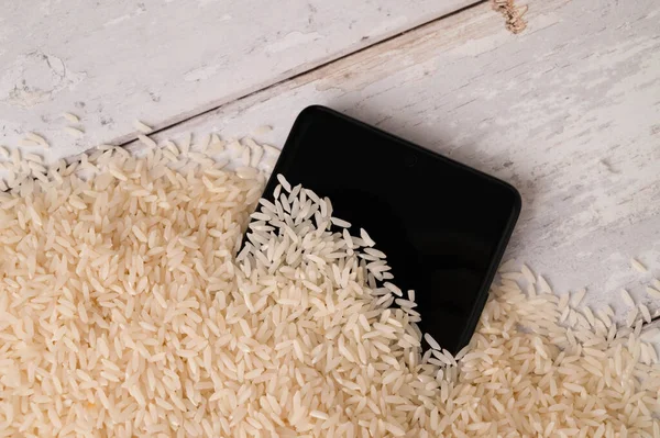 A close-up of a mobile phone placed to dry in a pile of rice after the phone fell into water. Light wooden table background. Top view.