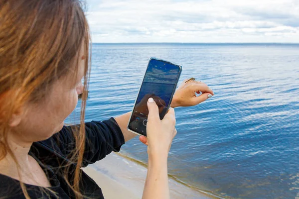 A girl photographs a dragonfly on the phone against the background of the sea close-up