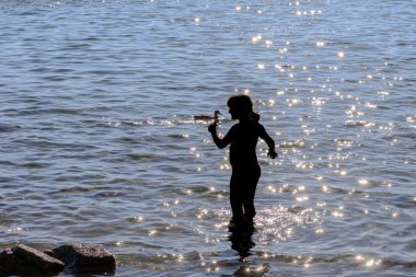 The silhouette of a child standing in the water and throwing water drops into the air is illuminated from behind by the setting sun