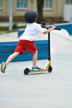 Çocuk scooter 'a biniyor. Scooter 'a binmek için oyun alanı. Beyaz tişörtlü, kasklı ve şortlu bir çocuk kaykay parkında scooter kullanıyor. Genç acemi sporcu boş zamanlarını ekstrem sporlarda geçiriyor..
