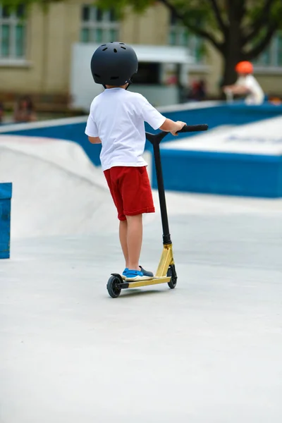 child rides a Kick scooter. playground for riding on Kick scooter. boy in a white T-shirt , hardhat and shorts riding a scooter in skatepark. young novice athlete spends free time in extreme sports.