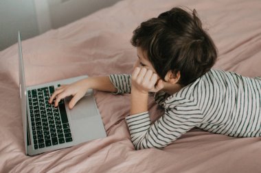 Küçük çocuk yatak odasında yatar ve dizüstü bilgisayara bakar.