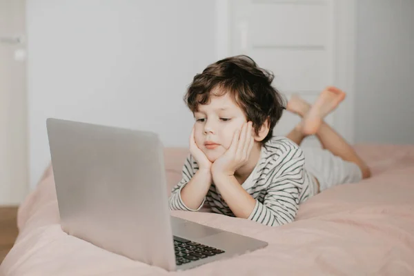 Маленький хлопчик лежить у спальні на ліжку і дивиться на ноутбук Стокова Картинка