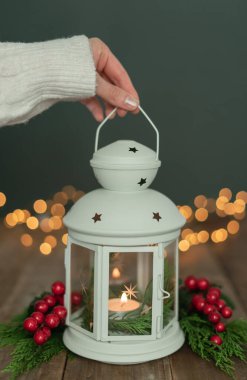 Beyaz kollu beyaz fener tutan, süslü bir kadın eli ve rustik ahşap masada Noel ışıkları. Boşluğu kopyala.