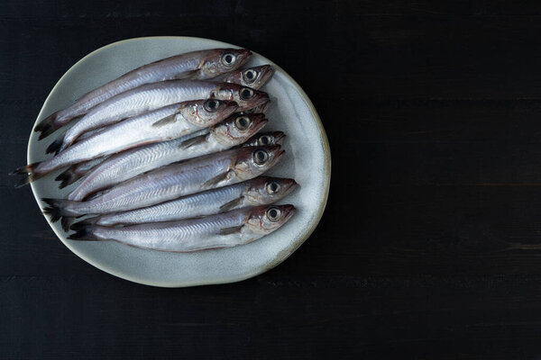 Fresh blue whiting on plate on black wooden background. Fresh fish. Copy space. Top view.