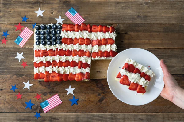 Square cake with the colors of the US flag on a wooden background with decoration. Hand with plate with slice of cake. Independence day celebration. Copy space. Top view.