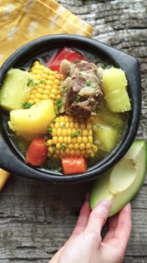 Typical Colombian Sancocho in a black ceramic bowl on a rustic wooden base. Womans hand placing avocado. Typical Latin American food.