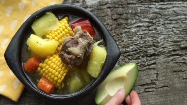 Blue plate full of sancocho on rustic wooden background. Hand placing avocado and putting coriander. Typical Latin American food.