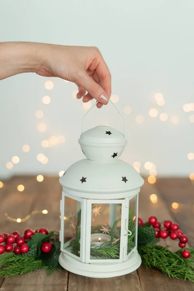 Woman's hand holding white Lantern with ornaments and Christmas lights on rustic wooden table. Copy space.