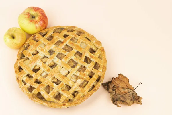 Baked apple pie with trellis decoration on a cream-colored background. Apples and dried leaves. Copy space. Top view.