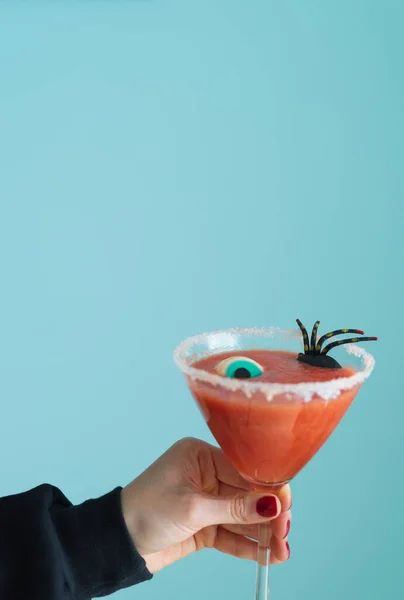 Woman's hand holds scary red cocktail with Halloween decoration on blue background. Vertical photo.