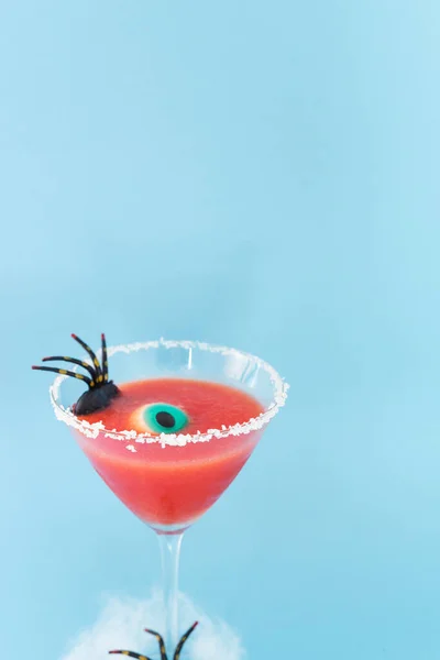 Scary red cocktail with Halloween decoration on blue background. Copy space. Vertical photo. Spider and eye decoration.
