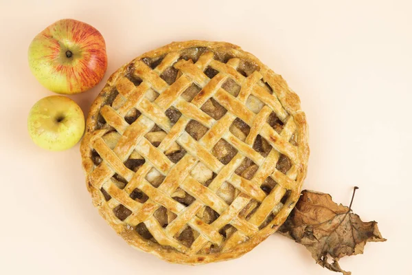Baked apple pie with trellis decoration on a cream-colored background. Apples and dried leaves. Copy space. Top view.