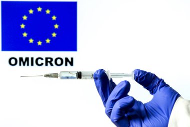 El ele tutuşma şırıngası ve covid aşısı. Arka planda yazılan Avrupa bayrağı ve Omicron covid değişkeni