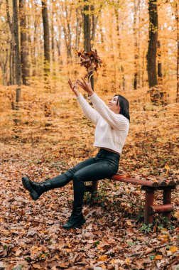 Güzel kız güler, zıplar ve sonbahar mevsiminde ormanda yapraklarla oynar..