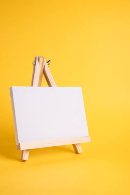 Küçük ahşap bir sehpada beyaz tuval sarı zeminde asgari konsept olarak duruyor. Boş boş çerçeve, kopyalama alanı olan tasarım için..