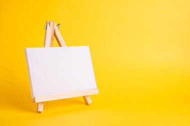Küçük ahşap bir sehpada beyaz tuval sarı zeminde asgari konsept olarak duruyor. Boş boş çerçeve, kopyalama alanı olan tasarım için..