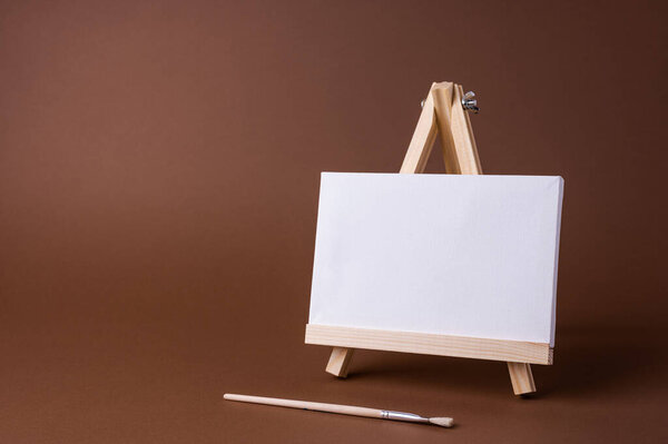 White canvas on a small wooden easel stand minimal concept on the brown backdrop. Empty blank mock up frame for design with copy space.