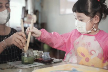Little Asian girl with facial hygienic mask and mother painting water colors together at home,selective focus. Quarantine