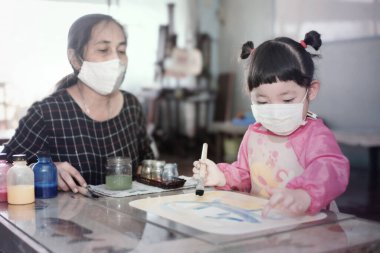 Little Asian girl with facial hygienic mask and mother painting water colors together at home,selective focus. Quarantine
