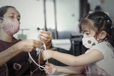 Little Asian girl wearing facial hygienic mask playing cats cradle game with her mother at home