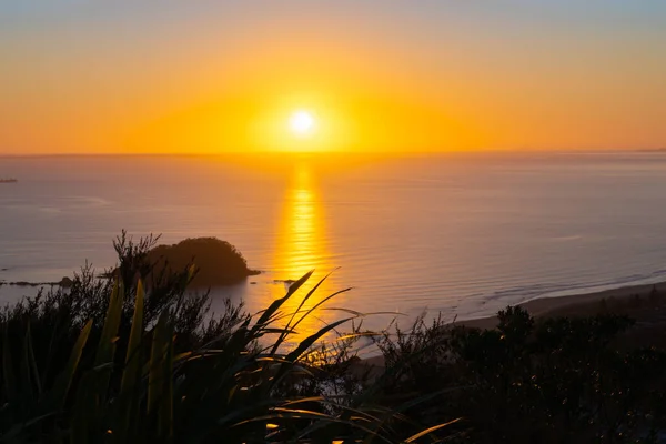 Golden glow of rising sun on distant horizon with long leading coastline of Bay of Plenty with Leisure Island from summit Mount maunganui.
