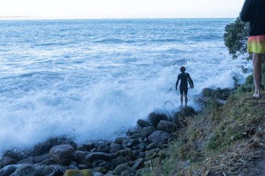 Tauranga Yeni Zelanda - 01 Ocak 2022; Genç erkek dalgalı deniz kenarında duruyor ve ne zaman atlama riskine gireceğine karar veriyor.