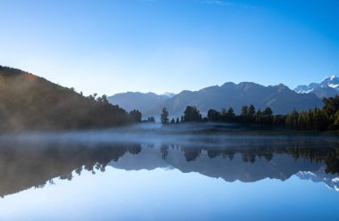Perfect reflection in Lake Matheson surrounded by beautiful natural forest under blue sky with view to Southern Alps and Mount Cook, South Island new Zealand. clipart