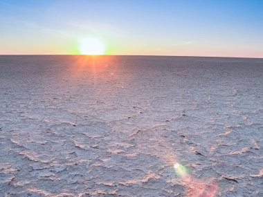 Glow of sun setting on horizon and lens flare across wide expanse of flat Makgadikgadi Desert Pan in Botswana.