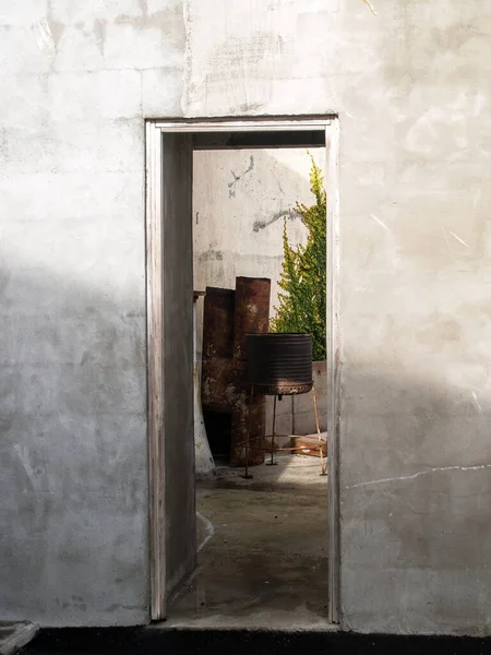 View through opening on concrete wall into courtyard with rusty old barbeque cooker and green ivy on wall.
