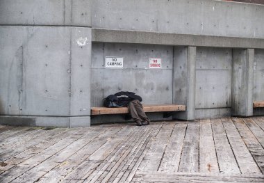 Poverty in city man asleep hiding under clothing on bench with signs above banning smoking and camping.