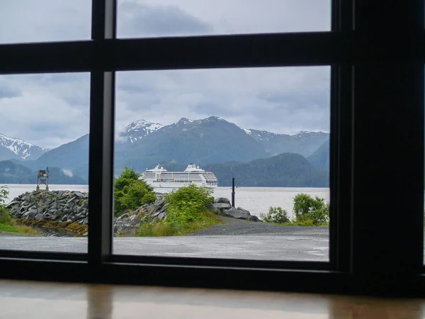 View through window of dramatic Alaskan scenery and passing cruise-ship under cloudy sky.