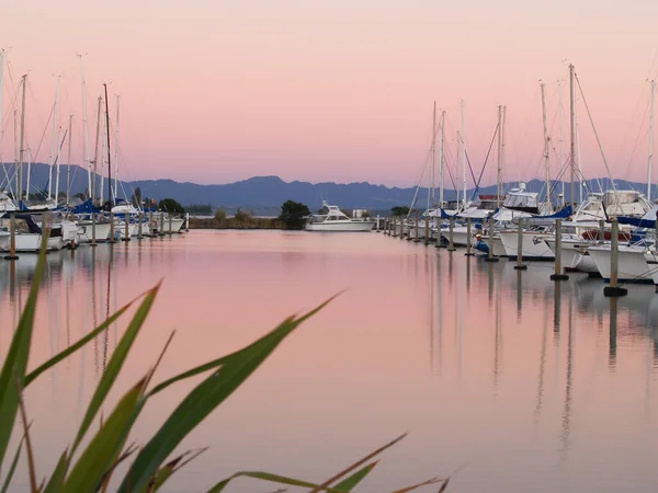 Boats moored in marina at sunset of beautiful pinks and blues reflected in calm water.