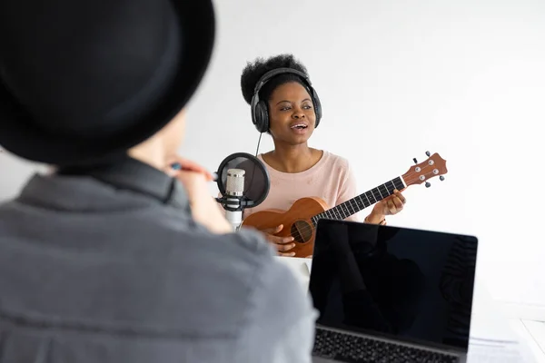 Grabar un podcast y crear contenido de audio. Una mujer afroamericana toca el ukelele en un estudio de grabación o en la radio — Foto de Stock