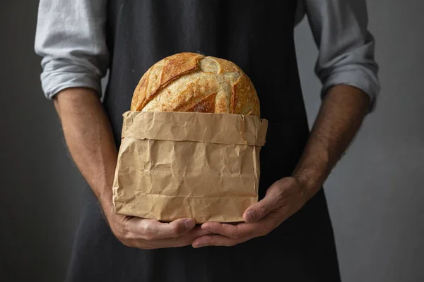 Egy felnőtt európai férfi pék kerek, friss kenyeret tart a kezében. egy pékségben lévő férfi élesztőmentes kenyeret tart kovászon és bagetten.. — Stock Fotó