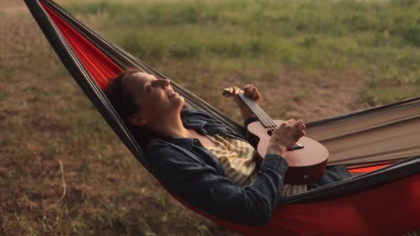 Uma mulher europeia em uma rede na natureza toca guitarra e canta. uma blogueira de viagens feminina cria conteúdo em vídeo — Vídeo de Stock