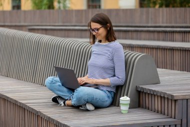 Genç ve kendine güvenen kadın blogcu ya da pazarlamacı kahve içiyor ve dizüstü bilgisayarı üzerinde çalışıyor. Parkta ya da halka açık bir yerde çalışmak. Sosyal medya ya da üst düzey yönetici olarak çalışmak.