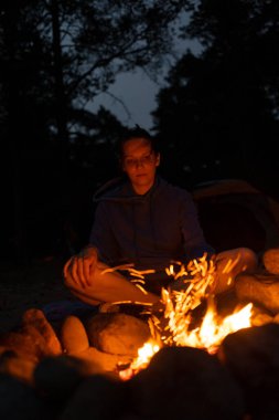 Bir kadın gece kamp ateşinin yanında oturur ve yürüyüş, yürüyüş ve açık hava aktivitelerinde ateşin arka planında çay içer.,