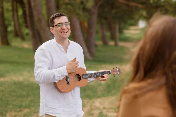 Um homem e uma mulher apaixonados abraçam-se e caminham na natureza. um homem toca guitarra ou ukulele para sua noiva ou esposa e se diverte — Fotografia de Stock