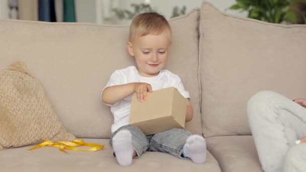Ένα μικρό αγόρι ξεπακετάρει ένα δώρο γενεθλίων ή Χριστουγέννων και χαίρεται. Ευρωπαϊκή οικογένεια μαμά και γιος δίνουν ο ένας στον άλλο δώρα για τα γενέθλια ή την ημέρα των μητέρων — Αρχείο Βίντεο