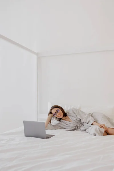 A European woman in a gray linen robe is working on a laptop at home or resting. a millennial woman is watching a movie on a laptop and having fun — Stockfoto