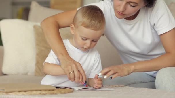 Η μαμά και ο γιος της ευρωπαϊκής οικογένειας ζωγραφίζουν με μολύβια και διαβάζουν ένα βιβλίο στο σπίτι. Η ανύπαντρη μητέρα μεγαλώνει ένα παιδί — Αρχείο Βίντεο