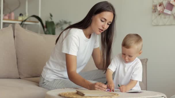 Η μαμά και ο γιος της ευρωπαϊκής οικογένειας ζωγραφίζουν με μολύβια και διαβάζουν ένα βιβλίο στο σπίτι. Η ανύπαντρη μητέρα μεγαλώνει ένα παιδί — Αρχείο Βίντεο