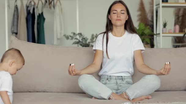 Una donna europea medita a casa sul divano. la madre di un bambino piccolo è impegnata in pratiche spirituali e yoga — Video Stock