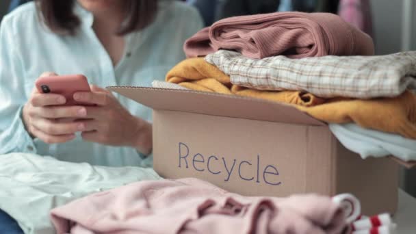 Una donna raccoglie vestiti in una scatola per il riciclaggio o lo smaltimento. una donna millenaria chiama il servizio di riciclaggio dal suo smartphone. — Video Stock